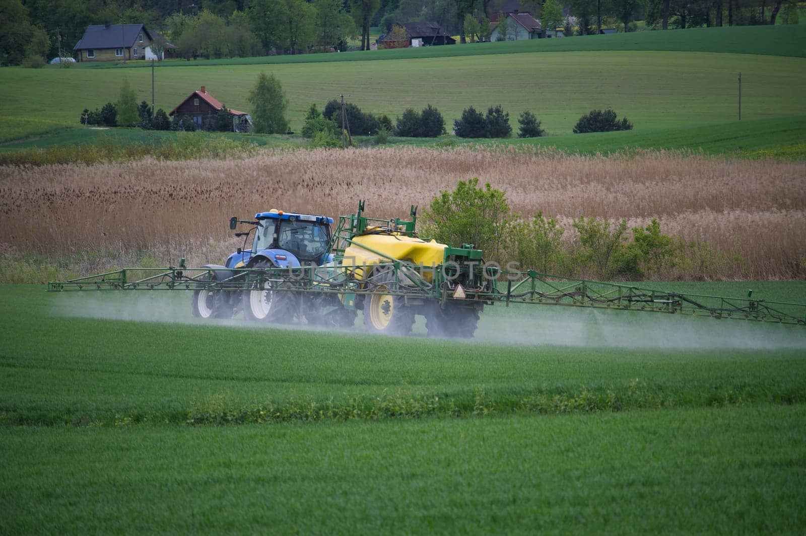 Farm tractor with sprayer disperses liquid over a lush green field amid a picturesque rural countryside setting. Surrounding the scene are farmhouse buildings, trees and vibrant agricultural landscape