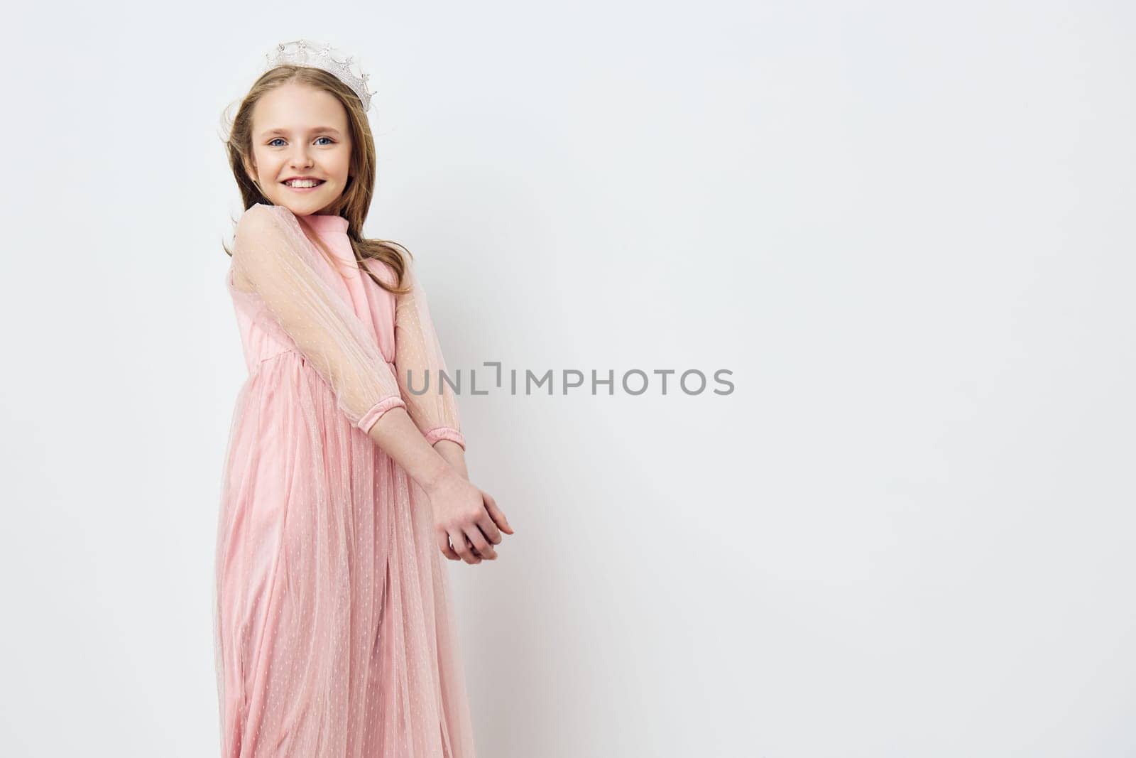 A cheerful young girl in a flowing pink dress with delicate sheer sleeves stands against a minimalist white backdrop, radiating happiness and playfulness, ideal for children s clothing, celebrations, and joyful moments