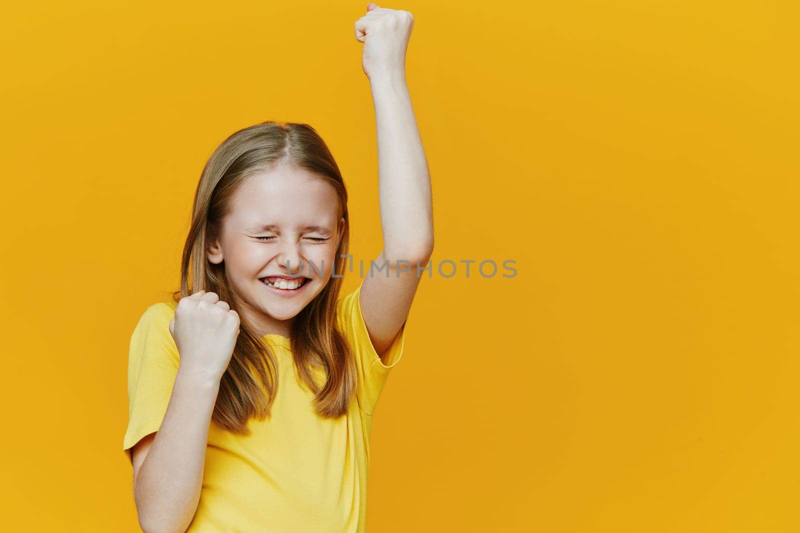 Joyful girl in a yellow shirt raising her fists in celebration against a bright yellow backdrop, embodying happiness and success Perfect for themes of achievement and positivity
