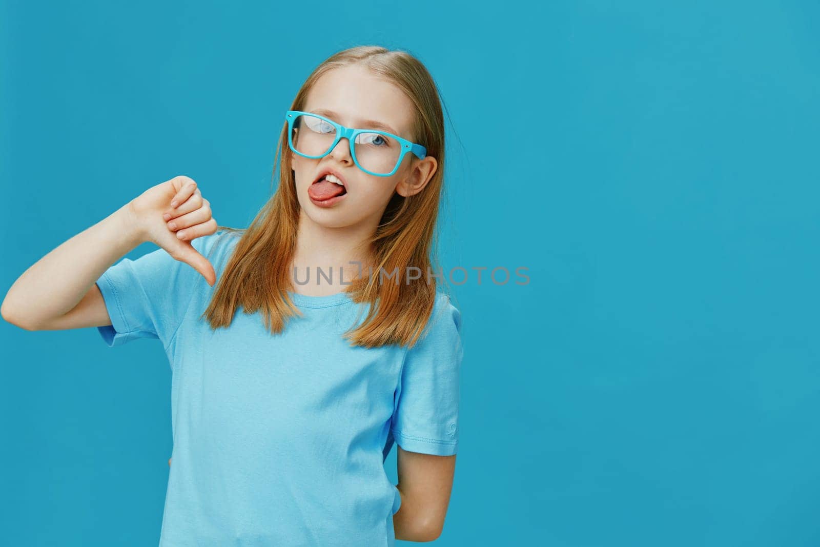 A playful young girl wearing blue glasses and a matching t shirt against a bright blue background, showcasing a fun and silly expression reflective of childhood joy