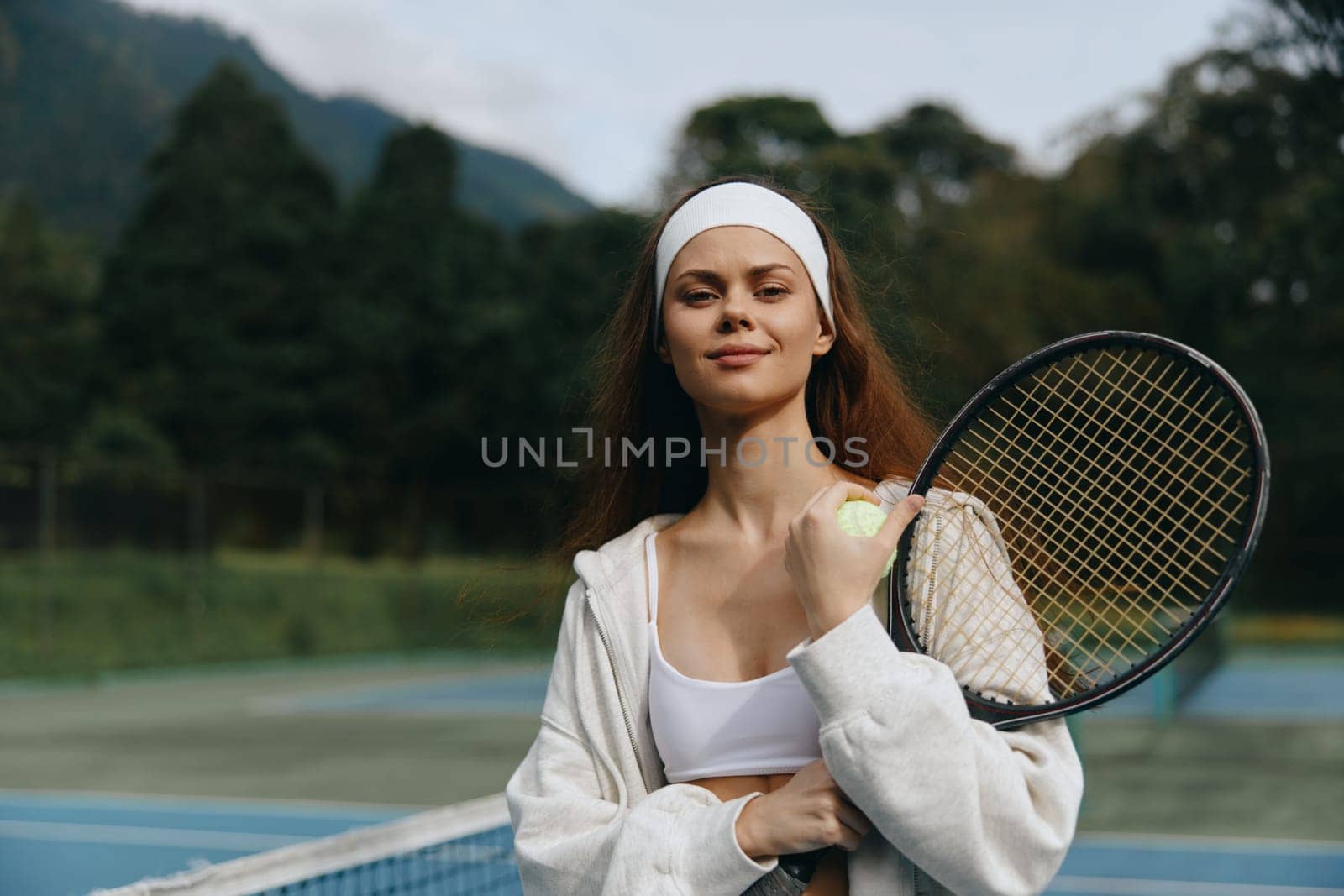 Confident young female tennis player in a sporty outfit and headband, smiling on the court with a picturesque nature background, embodying sportsmanship and fitness