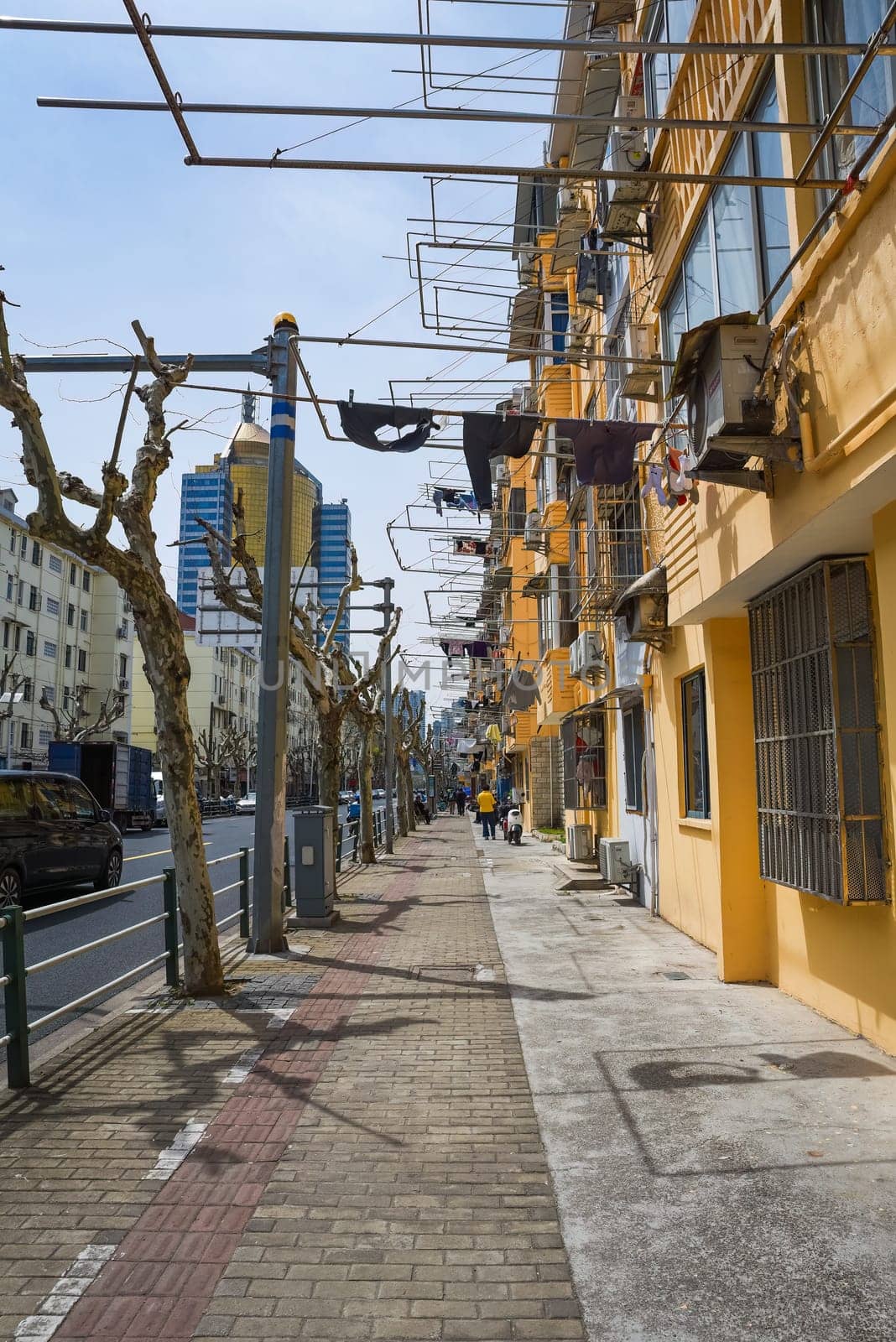 Shanghai, China - 1 April 2025: Traditional yellow apartments with laundry racks in Pudong. Pedestrians, trimmed trees, and a glass skyscraper under clear sky.