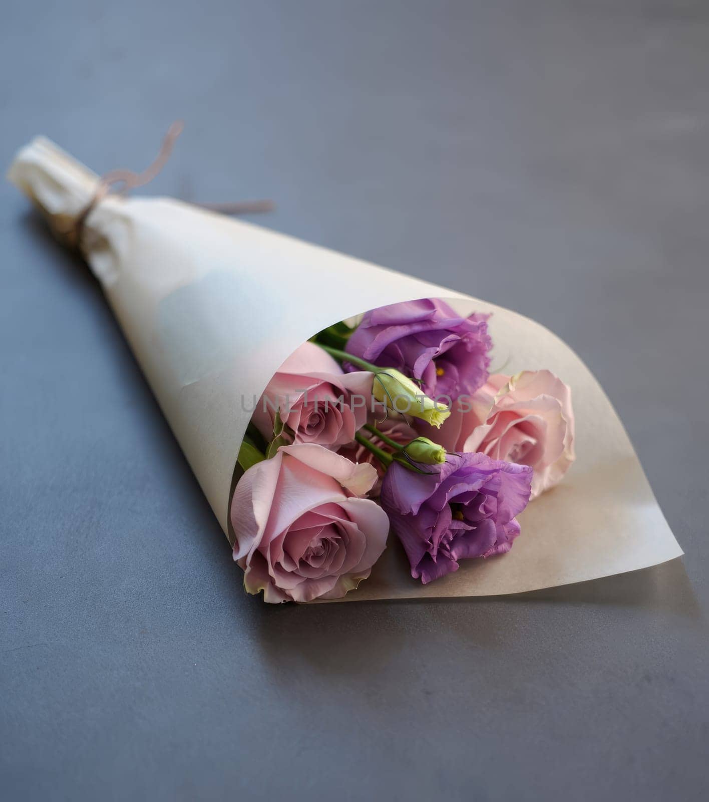 A bouquet featuring pink roses and purple flowers is elegantly wrapped in simple paper. This floral arrangement is placed on a smooth gray surface, highlighting its delicate colors.