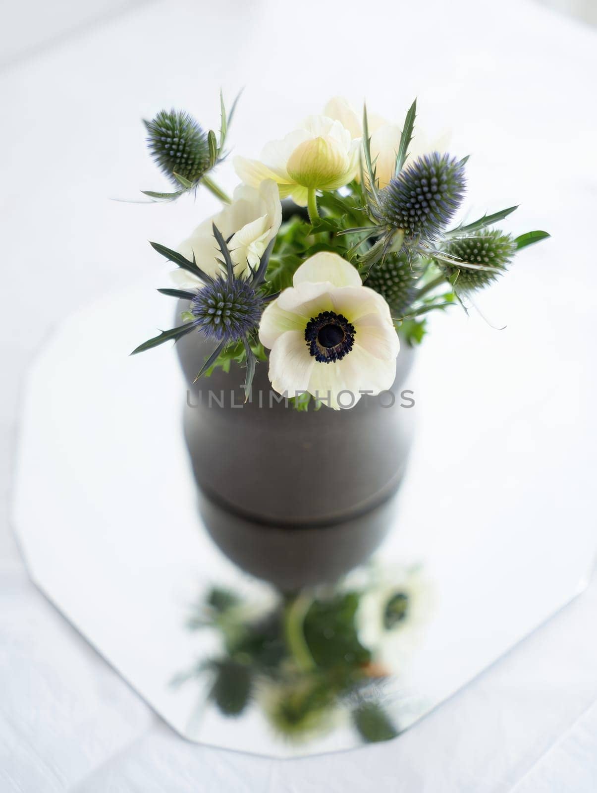 A delicate arrangement of white flowers and thistles sits in a simple vase on a smooth, reflective table surface, illuminated by gentle natural light.