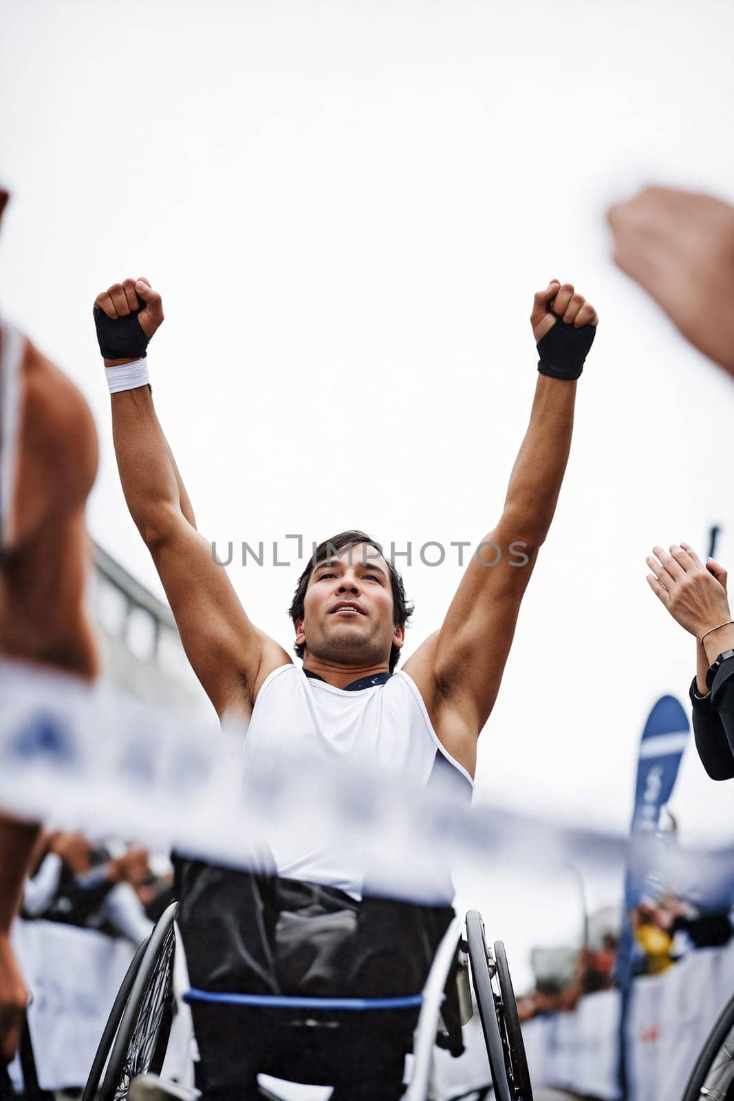 A determined wheelchair athlete raises his arms in triumph after crossing the finish line at a race. Spectators cheer in the background. Generative AI.
