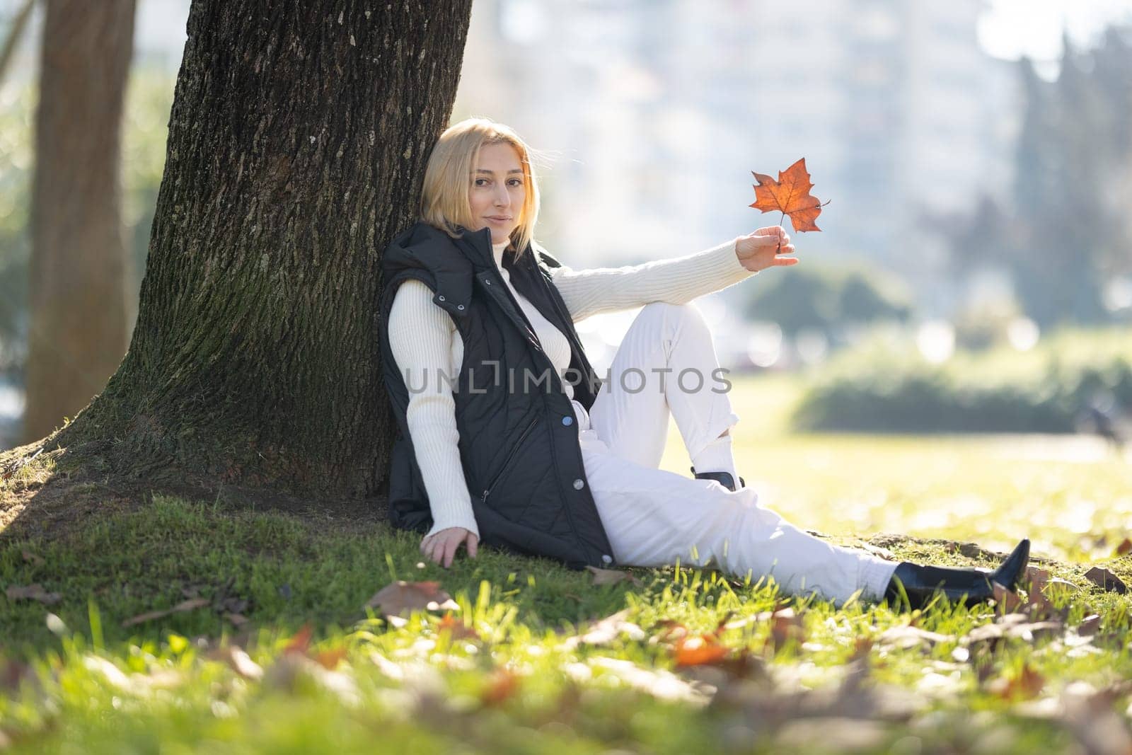 Young woman enjoying sunny autumn day in a park, holding a dry leaf and relaxing under a tree