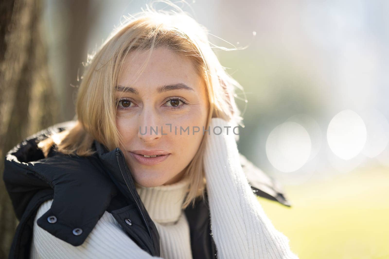 Young blonde woman wearing a white turtleneck sweater and a black vest, touching her hair while posing outdoors in a park during winter