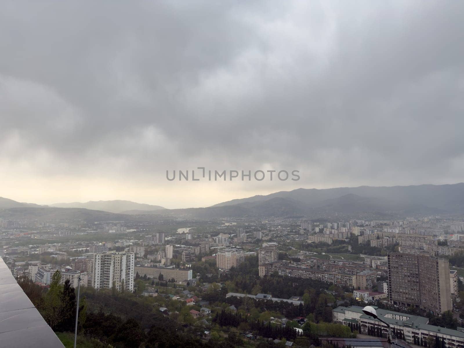 Tbilisi city view under dramatic sky surrounded by rolling hills of the Caucasus. Scenic perspective of Georgia's capital blending Soviet heritage and nature in atmospheric overcast weather