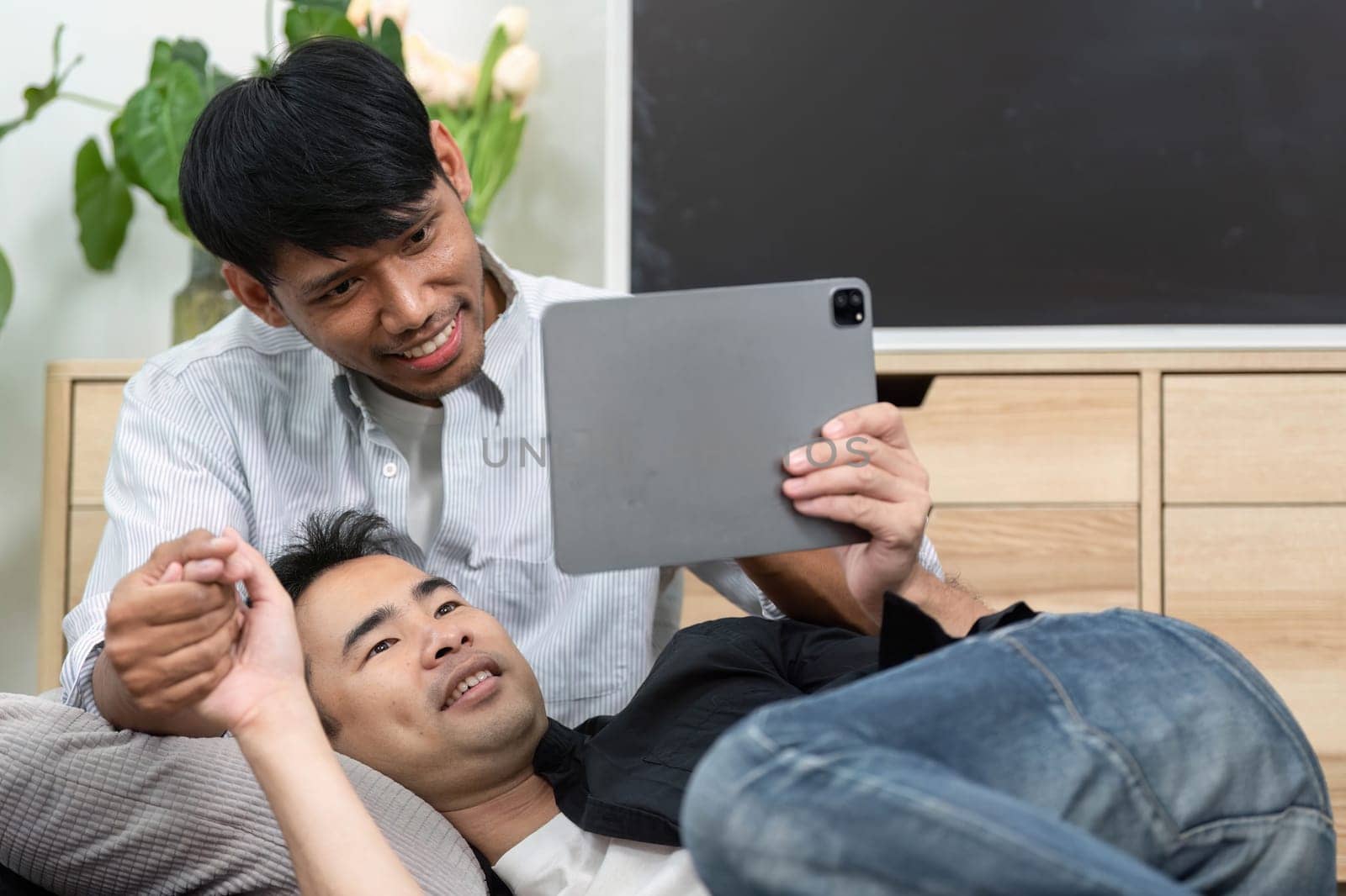 In a comfortable setting, a couple lies together, watching something on a tablet. Their interaction highlights the joy of shared experiences in an LGBT relationship, emphasizing love and connection.