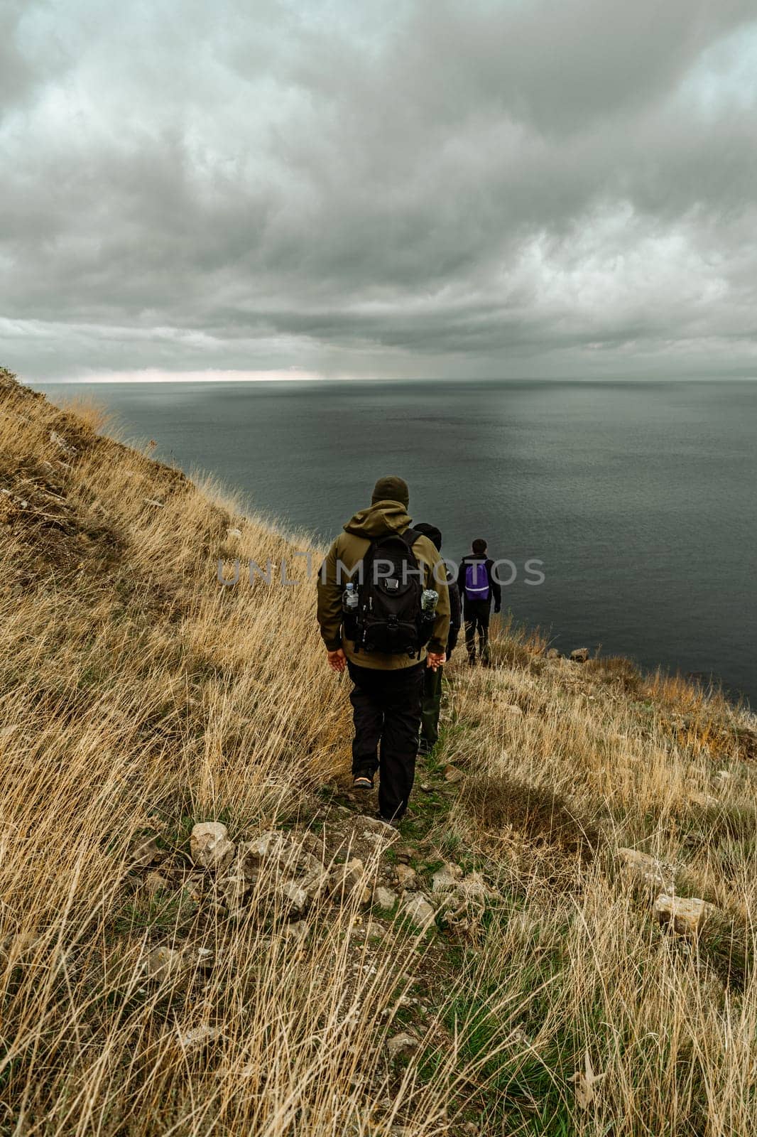 Three hikers descend a grassy slope toward the ocean, surrounded by rugged cliffs and shifting clouds. The atmosphere feels adventurous and serene as they navigate the landscape.