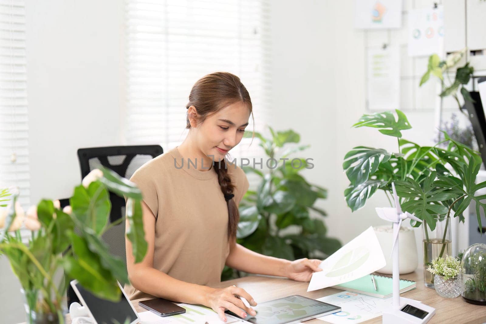 A young woman engages in sustainable work practices using her tablet and notes, surrounded by greenery in an eco-friendly office space.