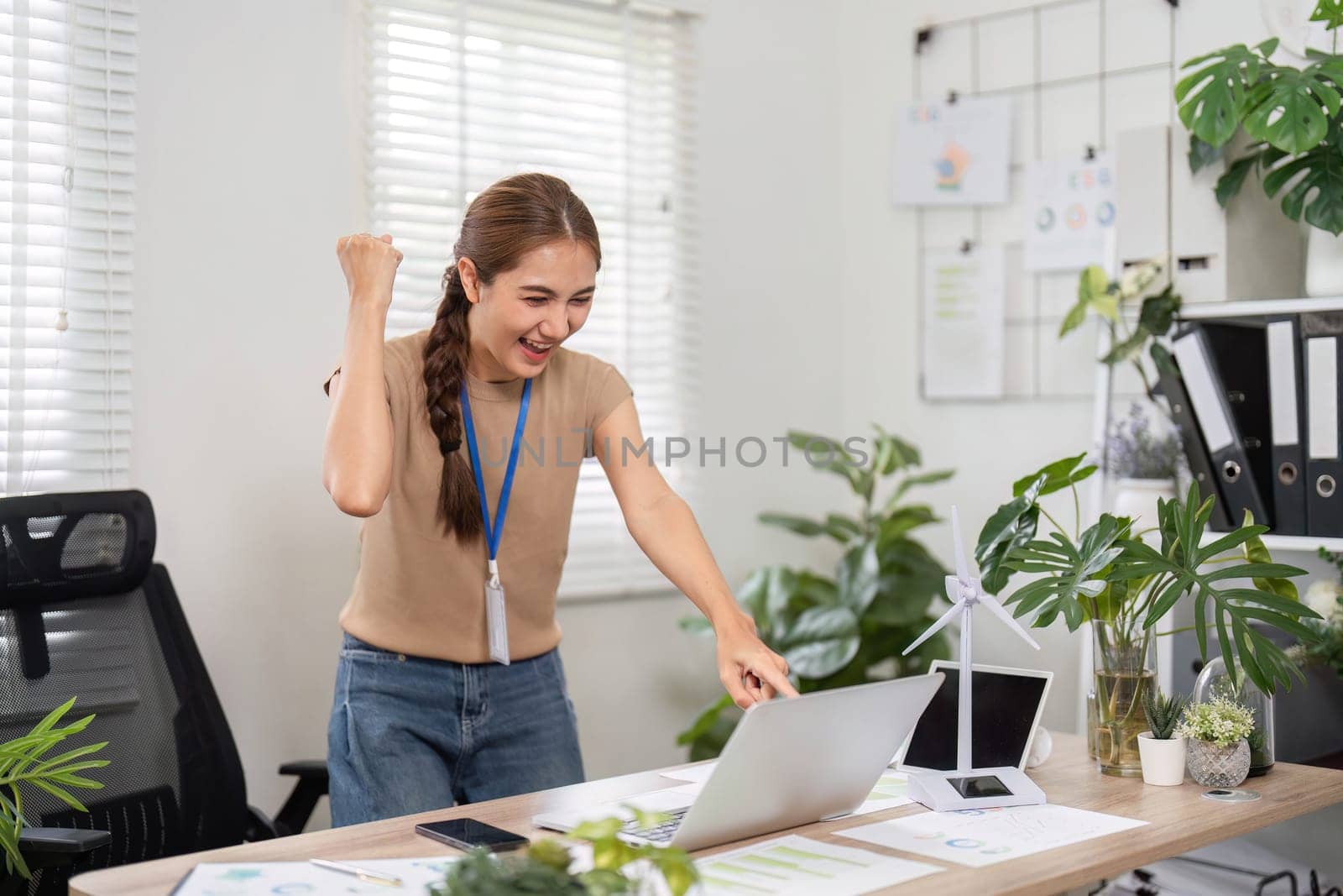 A young woman at her office desk, celebrating a recent achievement, surrounded by plants, showcasing a vibrant and sustainable work environment.