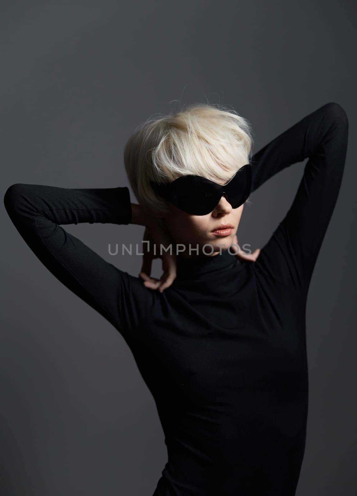 Confident young female model with short platinum hair and stylish sunglasses showcasing a modern and chic fashion look, set against a simple dark gray backdrop.