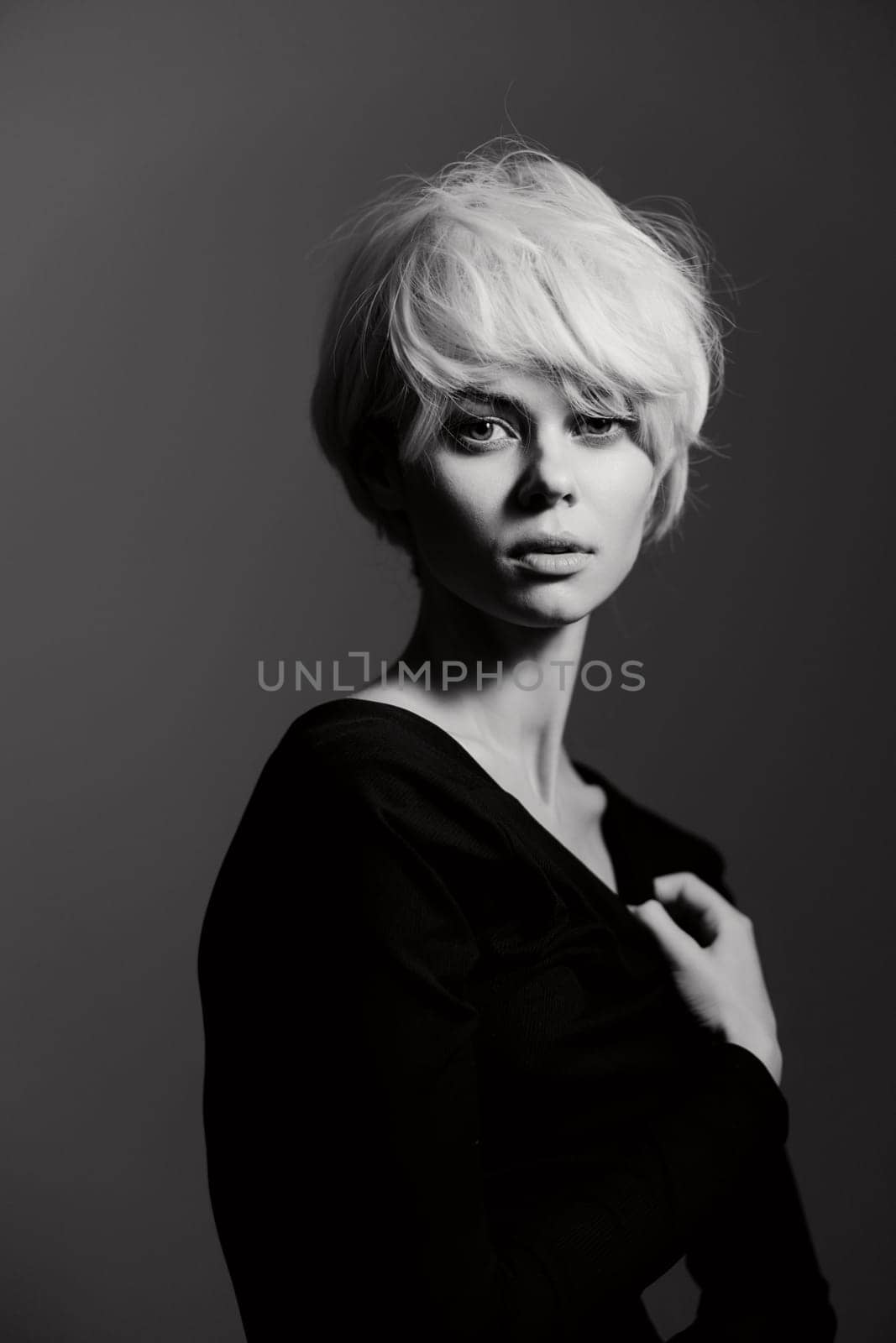 A striking black and white image featuring a young woman with short, tousled blonde hair and a black off-shoulder top. Her thoughtful expression contrasts with the soft neutral background, creating a