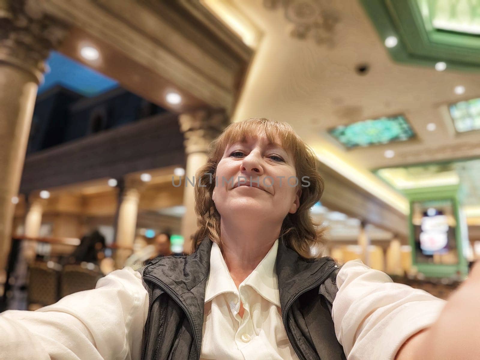 A middle aged blonde woman enjoys her travel experience while capturing a selfie at a stunning venue filled with vibrant decor.