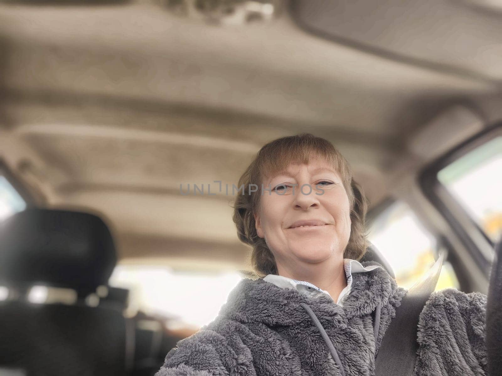 A middle-aged woman captures a cheerful moment while driving in her car, dressed comfortably in a soft hoodie.