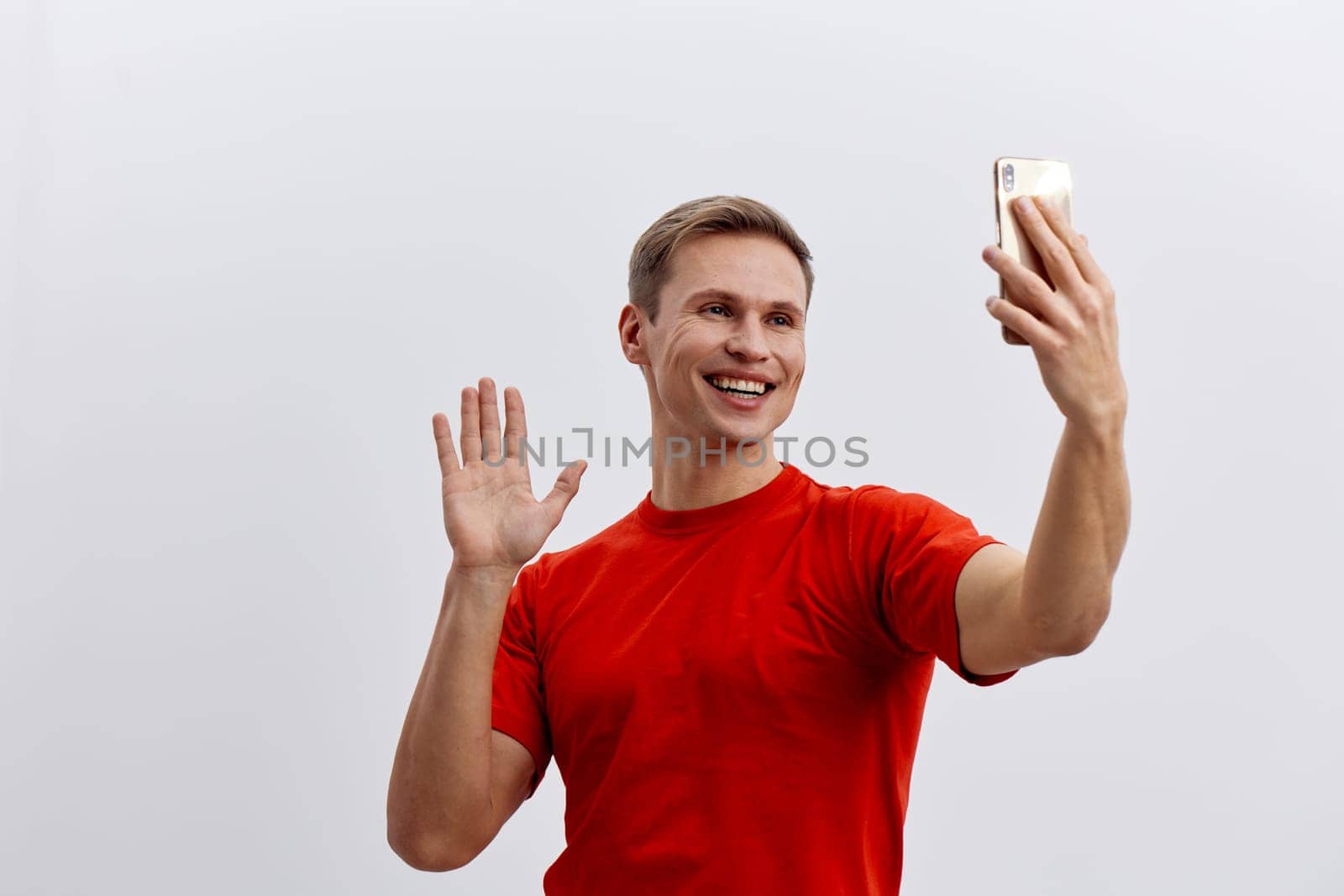 Smiling man in casual red t-shirt capturing selfie with smartphone, cheerful expression, waving hand, modern lifestyle, bright indoor setting, isolated on plain light background, concept of technology and happiness