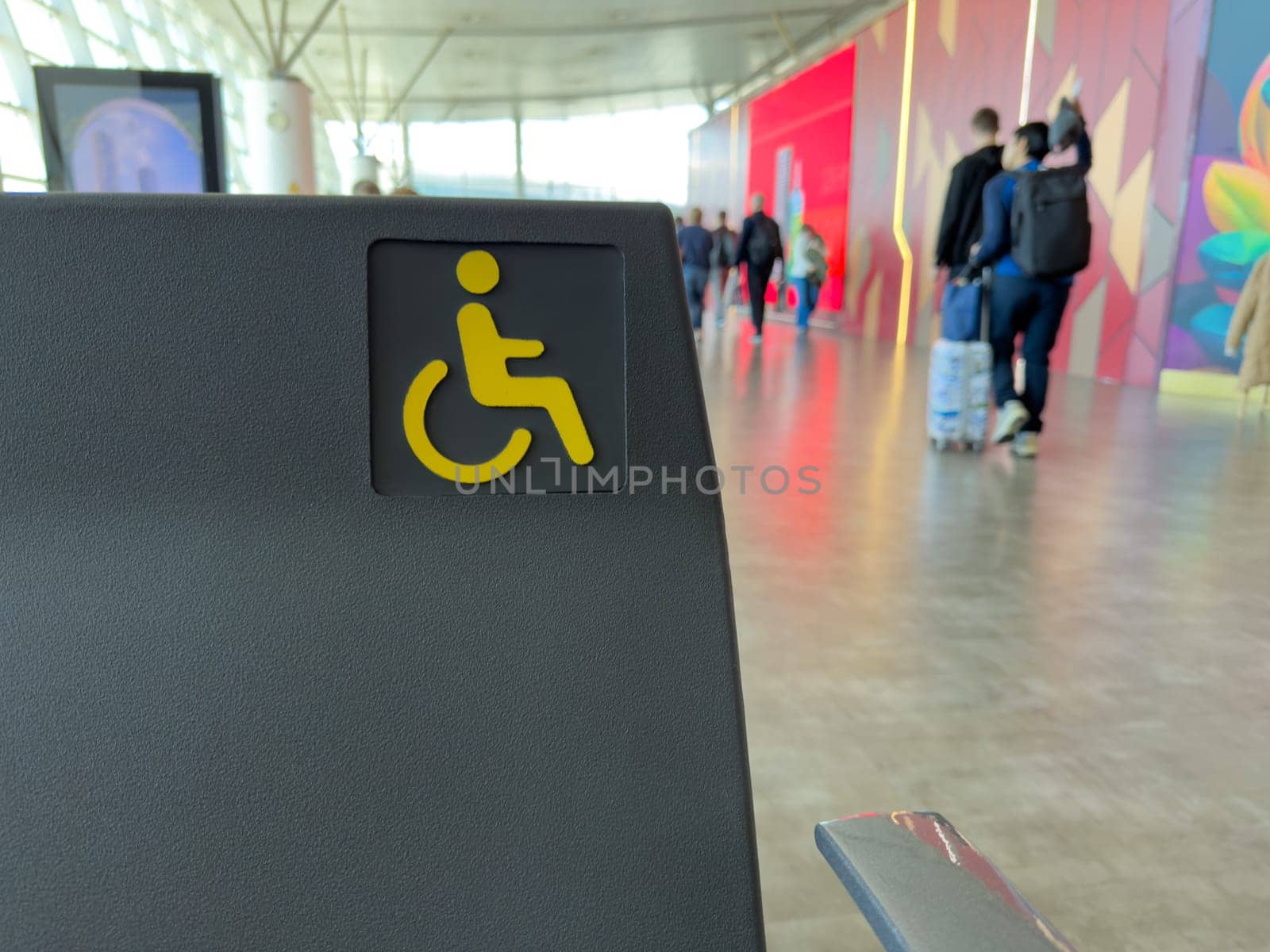 Accessible seat marked with yellow wheelchair icon in public indoor space, with people walking in background, inclusive travel and accessibility in transportation hubs