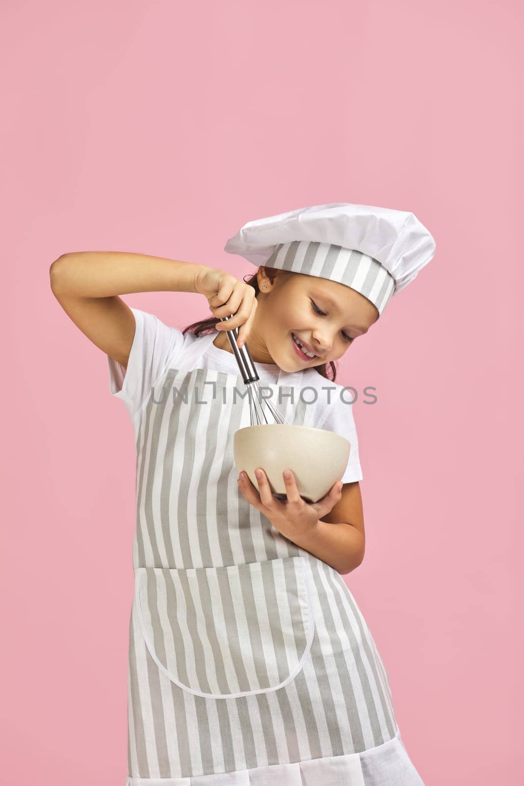 smiling little girl in chef hat and apron mixing something with a whisk in bowl over pink background. child dreams of becoming a chef