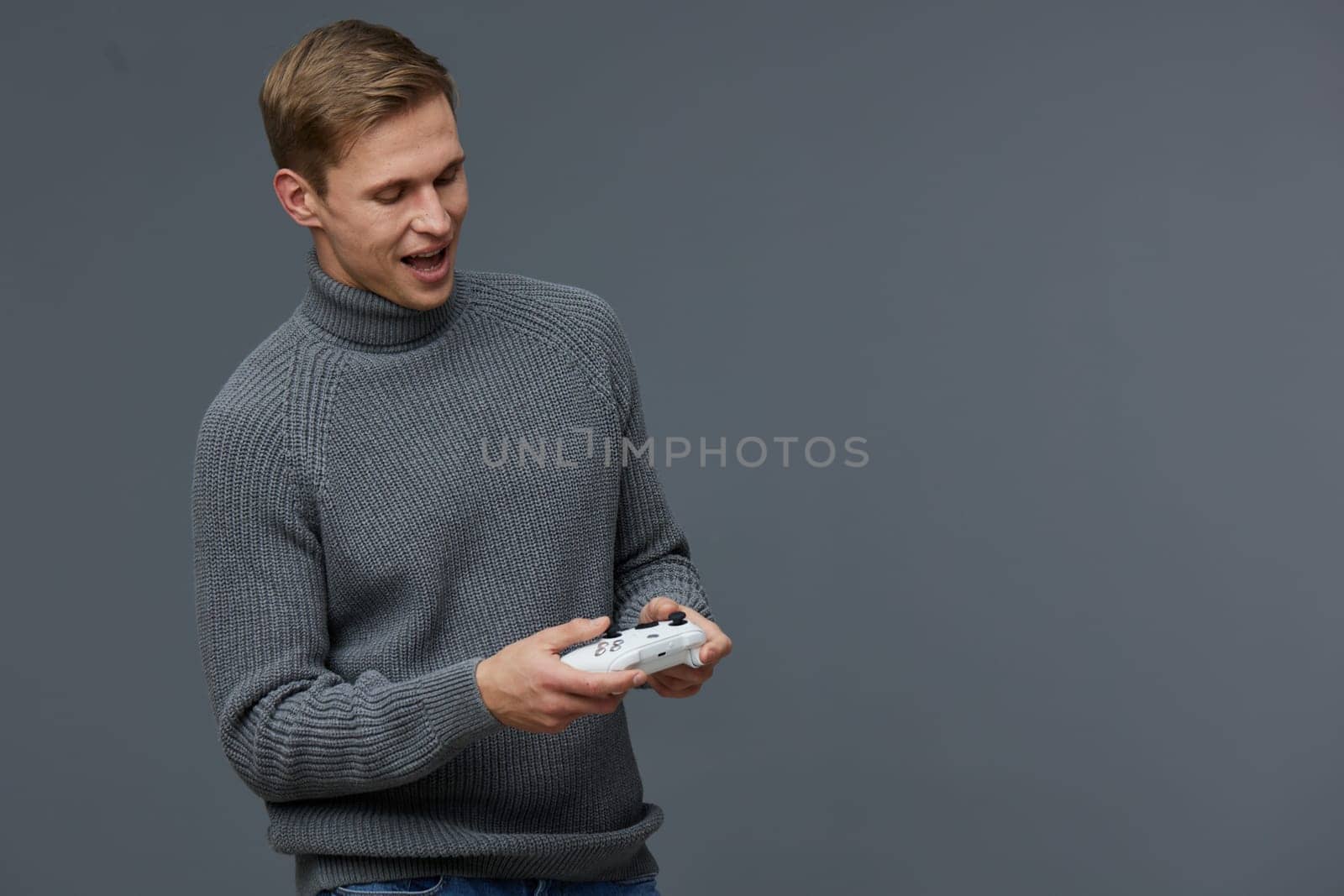 Happy young man in gray sweater playing video games with game controller, standing against a neutral gray background. The image reflects leisure, entertainment, and modern technology, capturing a cheerful and relaxed mood. Concept: gaming enjoyment.