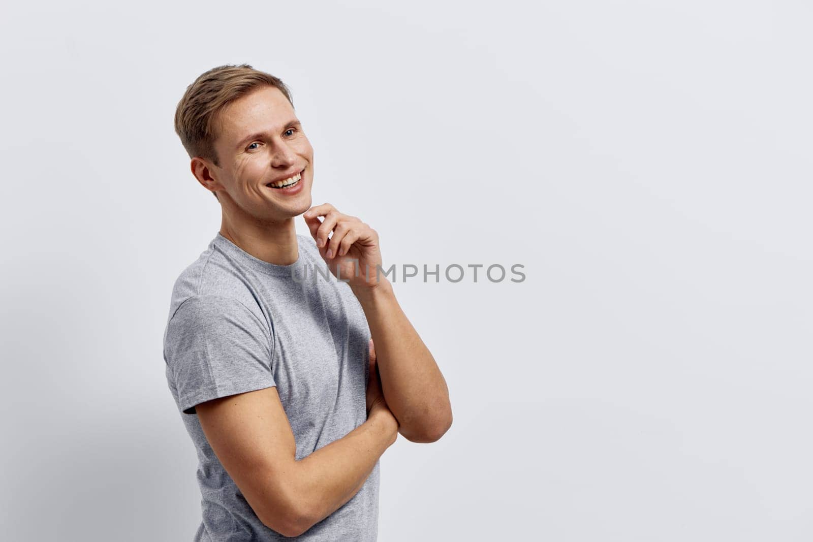 Smiling young caucasian man in casual grey t-shirt stands with arms crossed looking aside with thoughtful expression against plain light grey background. Concept of digital nomad working remotely with technology and innovation.