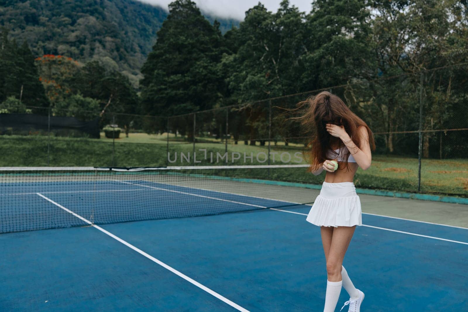 A youthful tennis player in a white outfit on a blue court, joyfully holding a ball, amidst green scenery and mountains, capturing the spirit of the game and summer