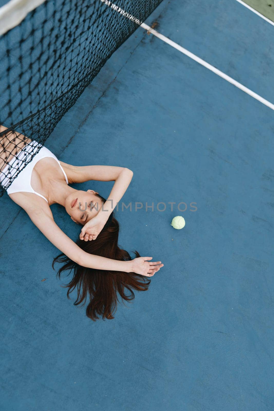 A serene young woman in a white outfit lies on a blue tennis court, symbolizing a sports lifestyle and emphasizing relaxation and wellness in sports activities