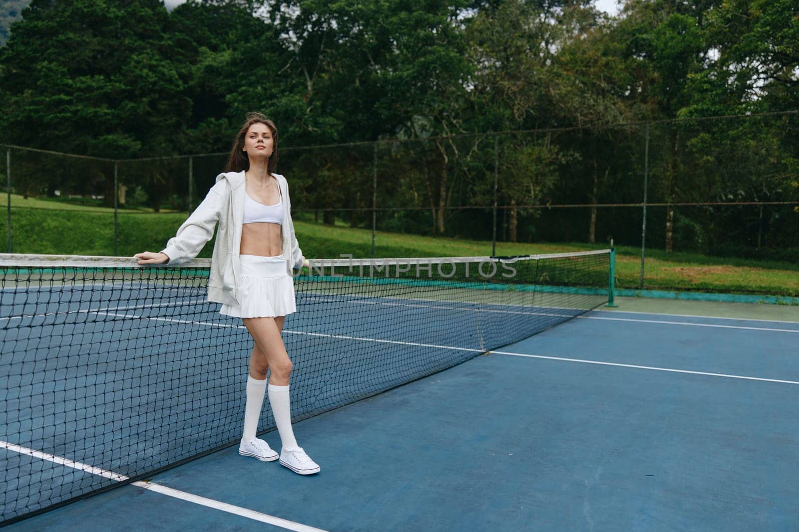 Confident young woman in white sports attire poses by the tennis net, showcasing a healthy lifestyle and sports enthusiasm, ideal for fitness and sporting themes