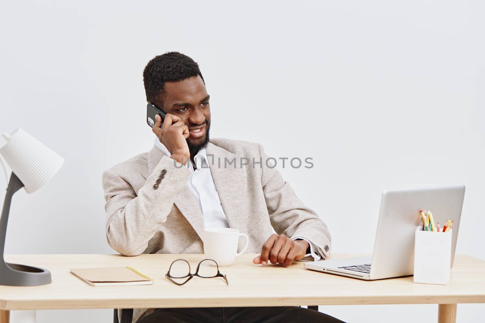Business professional man with joyful expression speaking on cell phone while seated at desk in contemporary office space, surrounded by laptop, glasses, and paperwork, conveying communication, workplace, success, and technology concepts.