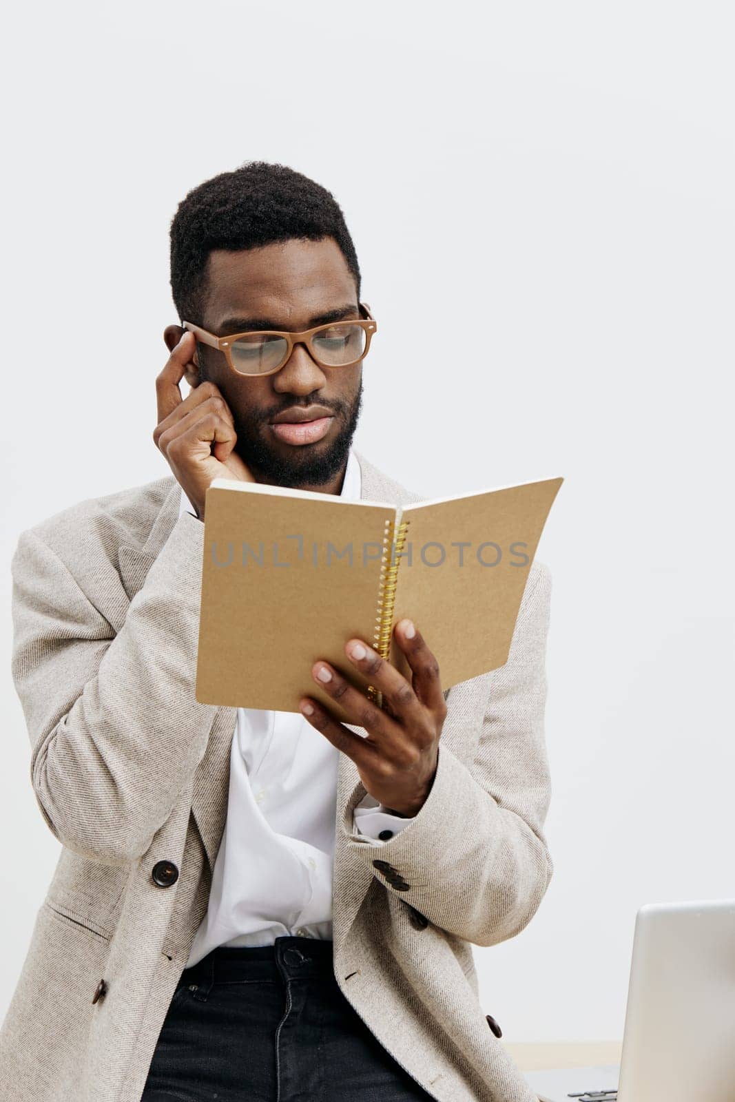 Focused young man of African descent with glasses, dressed professionally in a beige blazer and white shirt, reading a notebook attentively, sitting at a desk with a laptop, plain white background, indoor work environment. Concept: workplace concentration