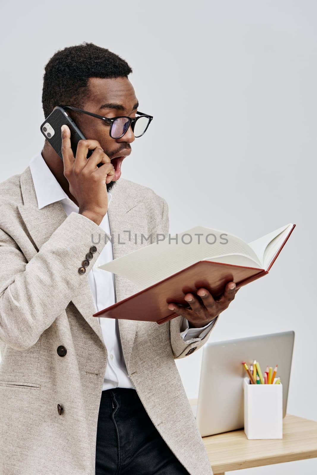 Confident young man of African ethnicity with glasses, dressed in a beige blazer and white shirt, engaged in phone conversation while holding an open notebook, standing in a clean, minimalistic office with a desk and stationery. Office worker lifestyle