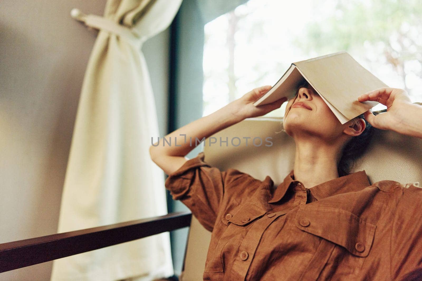 Young woman of Asian descent relaxing in a serene indoor environment, using a book to shield her face from light The warm color palette enhances the cozy atmosphere