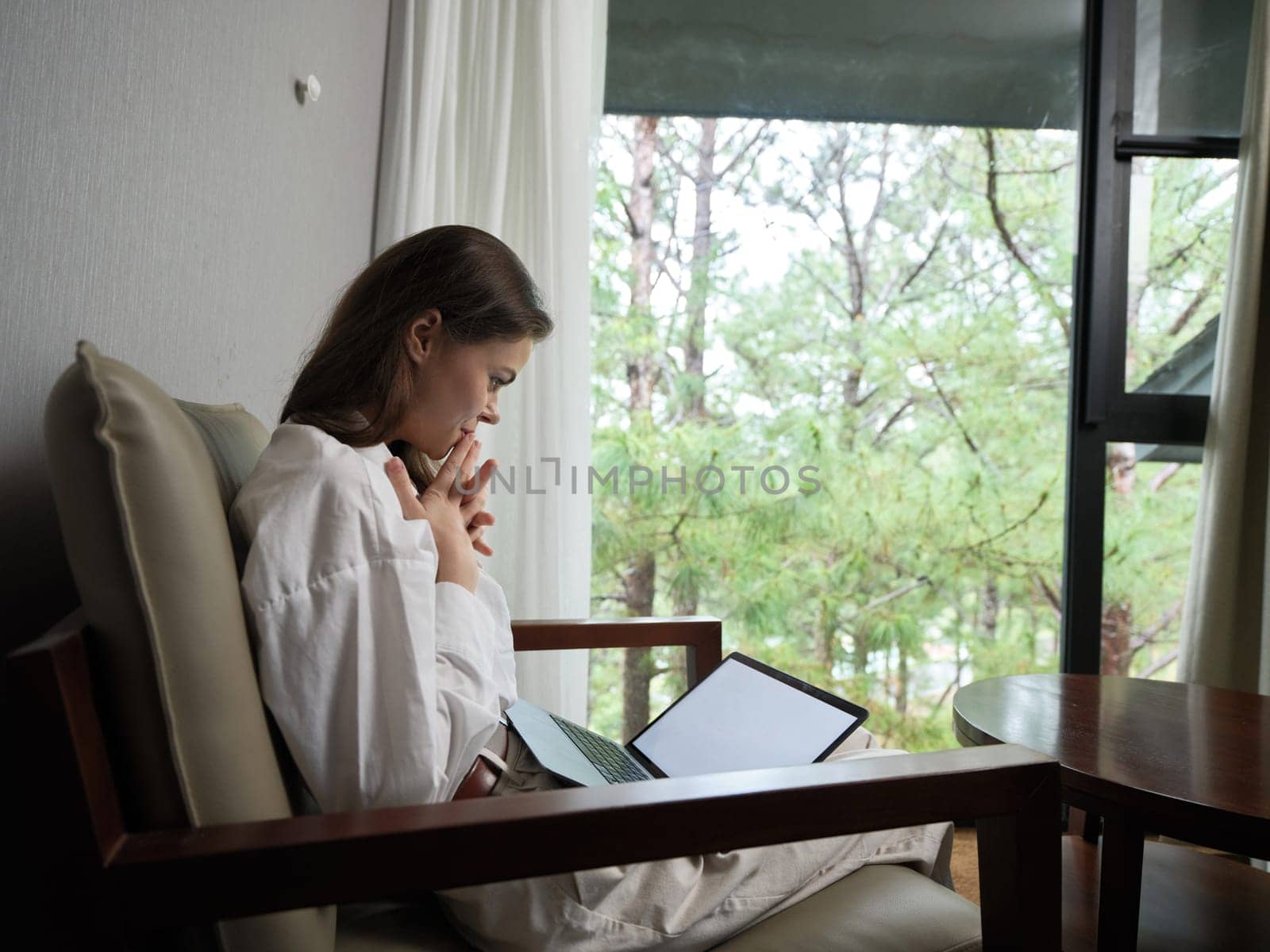 A young woman in a white outfit sits by a large window, concentrating deeply as she works on her laptop, surrounded by a peaceful green nature backdrop