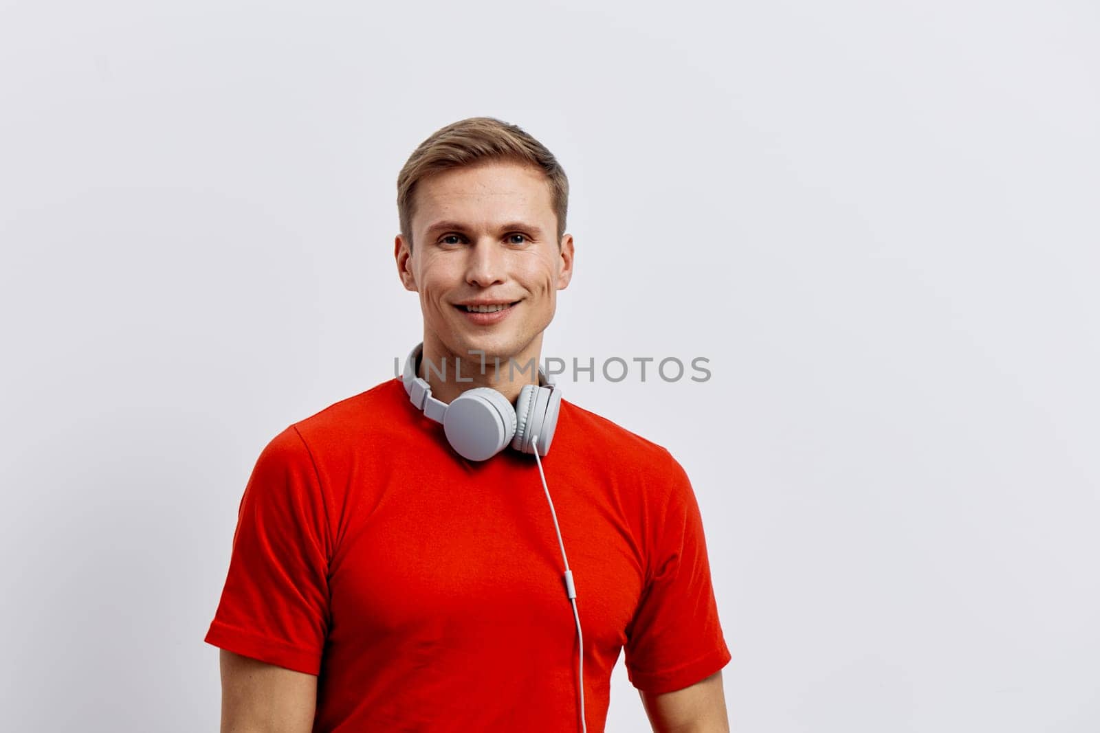 Happy young man with short blond hair, dressed in a bright red t-shirt, is smiling and listening to music through white headphones while posing in a studio with a white background. Concept: enjoying music and leisure.