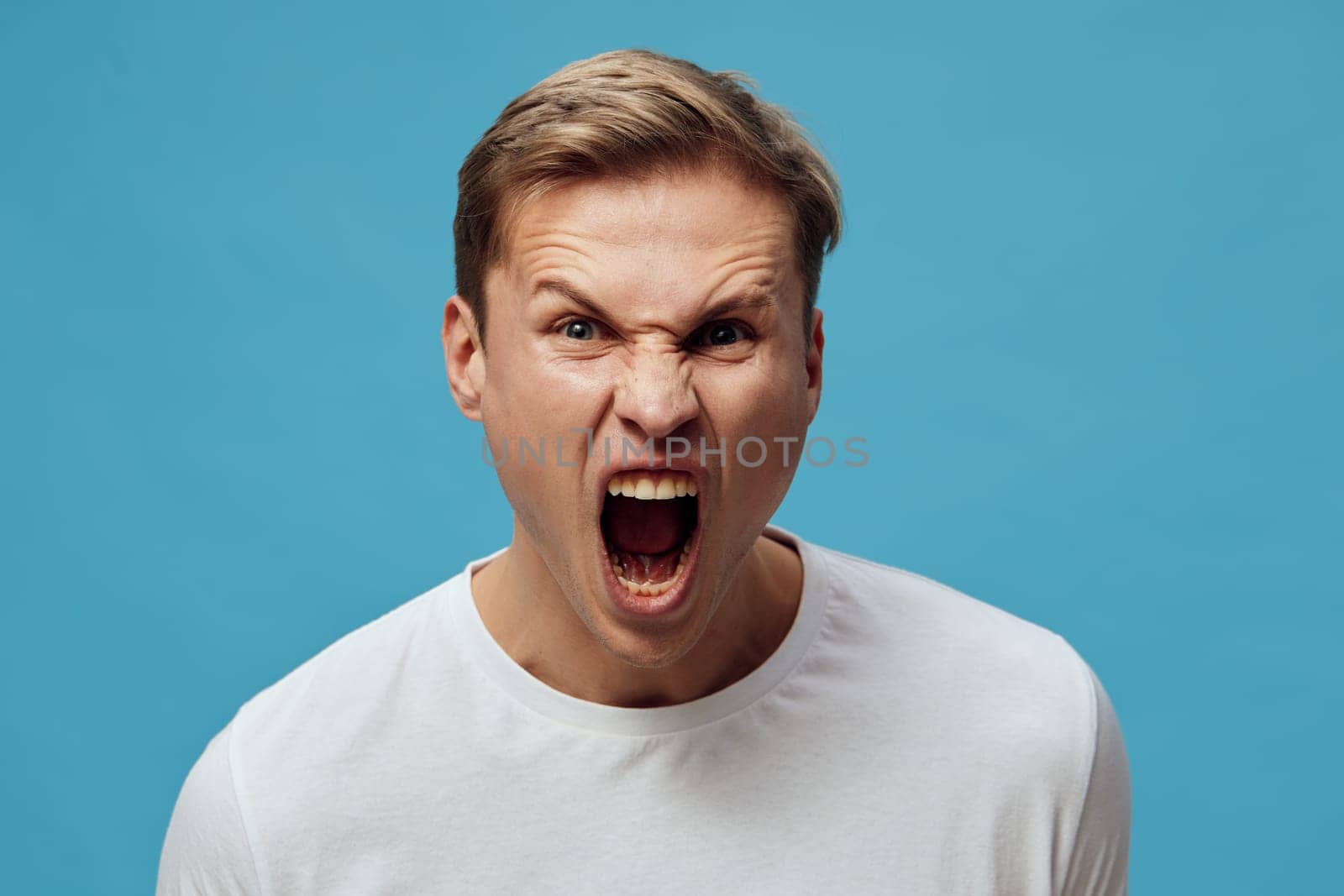 Portrait of frustrated young man with short brown hair, expressing anger or rage through shouting with open mouth, wearing casual white t-shirt, against plain blue background. People emotion concept