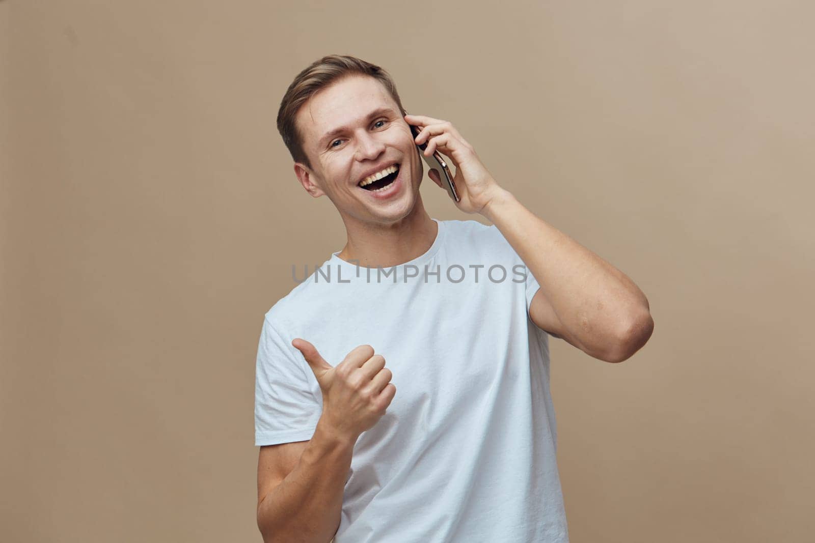 Cheerful smiling man engaging in a phone call on mobile device with a thumbs-up gesture, casual attire, light brown hair, in a neutral beige background. Modern lifestyle and communication concept