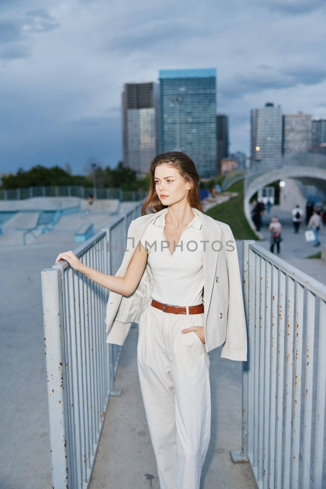 business woman in white outfit wearing blazer standing outdoors by metal fence with modern cityscape background, conveying confidence and professionalism