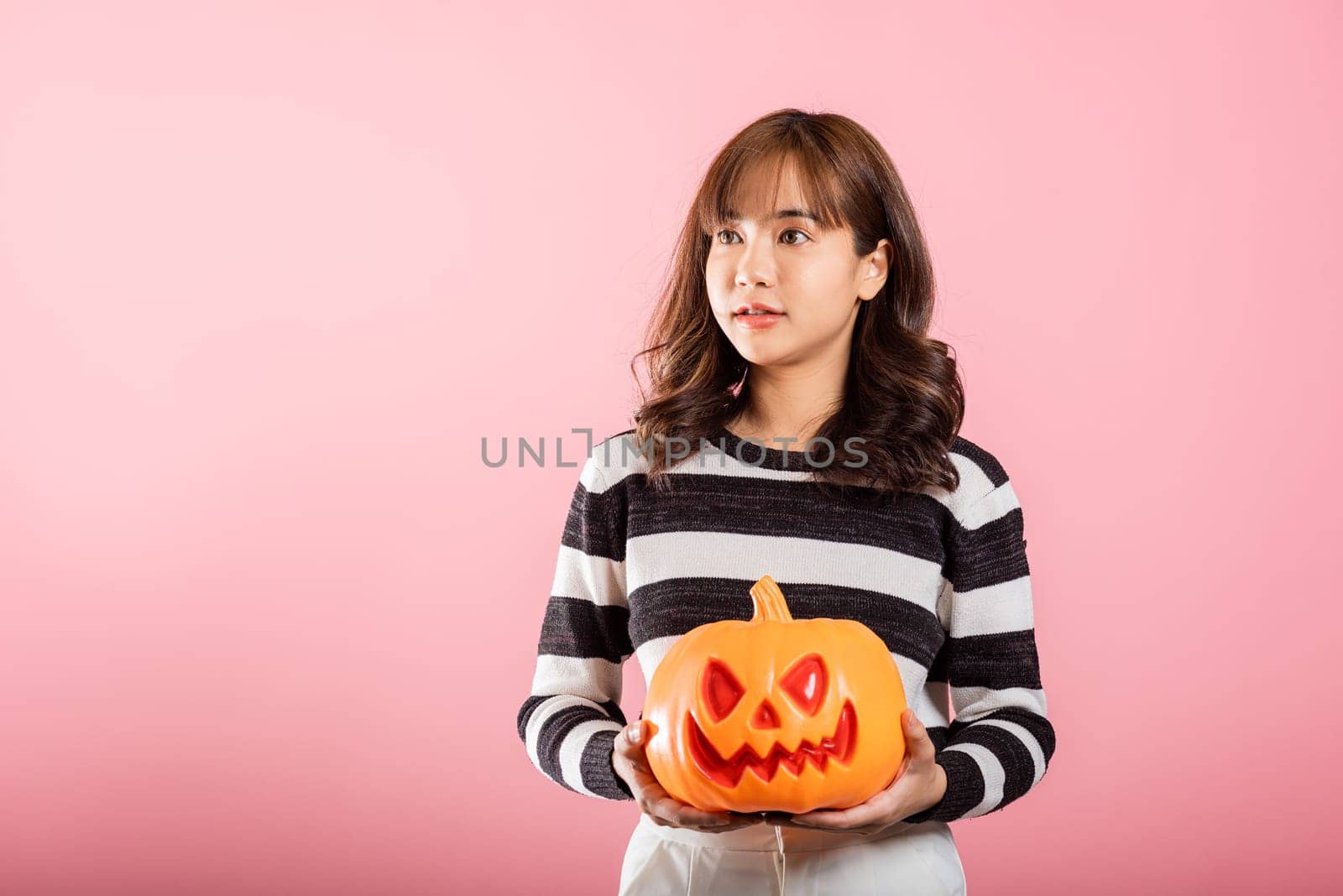 Halloween-themed studio shot, an Asian woman holds a variety of model pumpkins, including a ghost-shaped one, on a pink background. Her cheerful costume sets the tone for a fun and spooky Halloween.