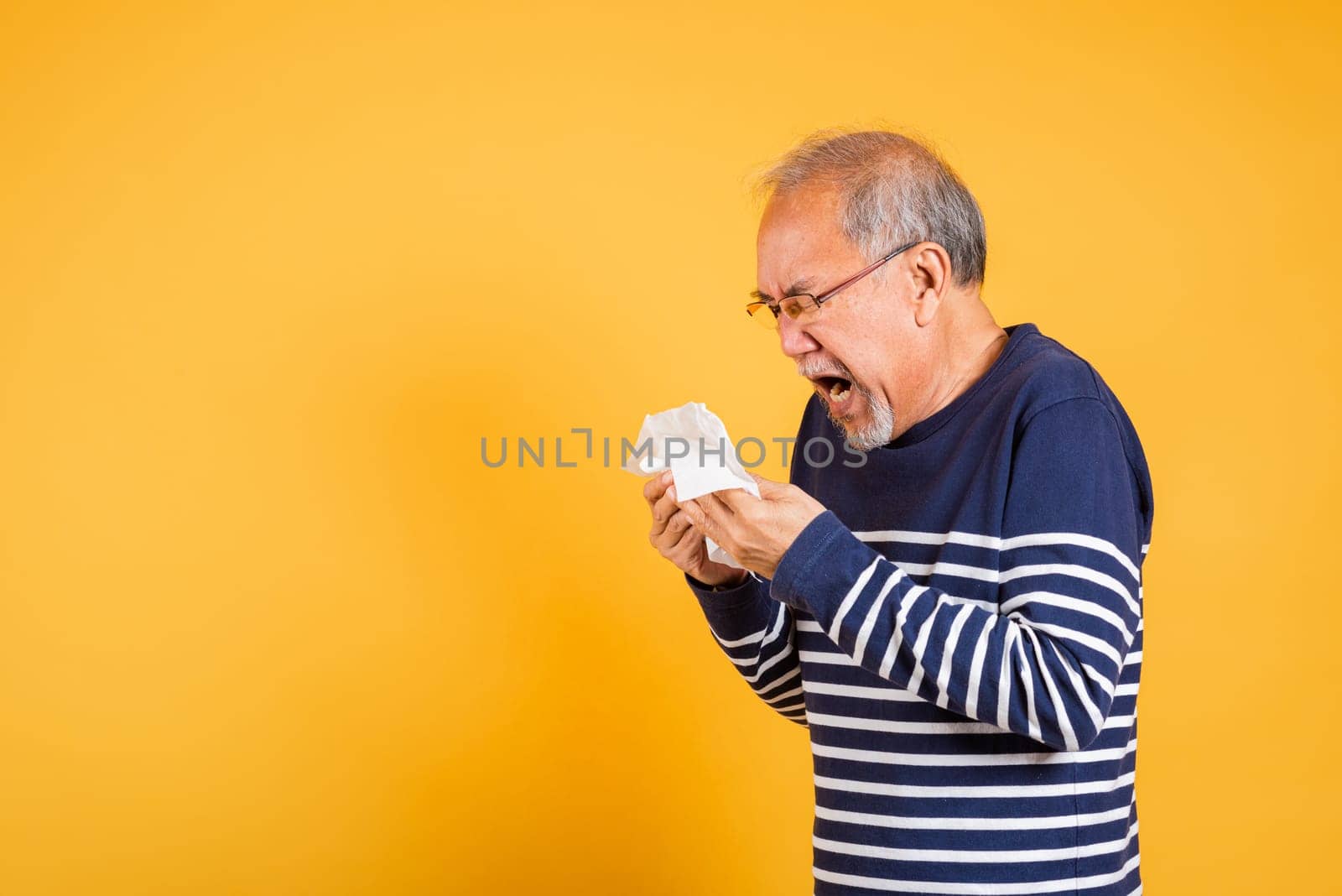 Portrait senior old man blowing nose with tissue studio shot isolated on yellow background, Asian elder man cold having flu and sneezing from sickness virus problem use tissue, pensioner unwell