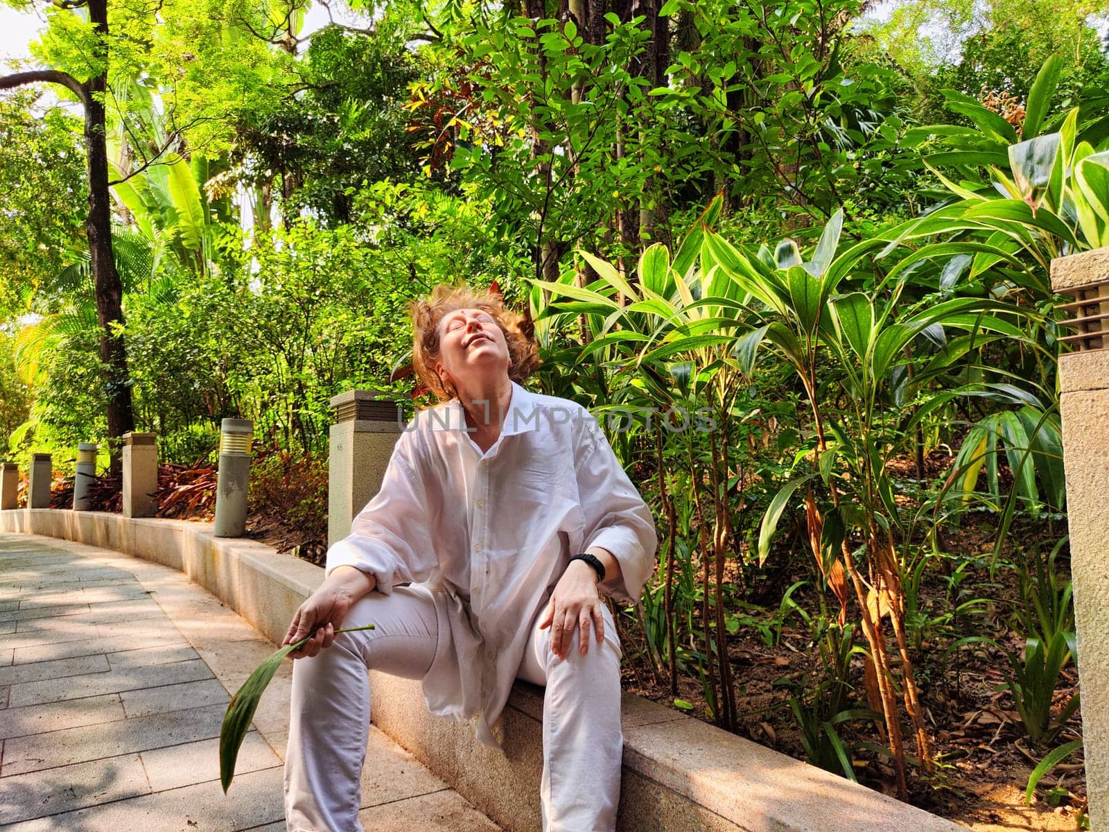 Blonde woman relaxes amid vibrant greenery, soaking in the sun during a leisurely outdoor adventure in a scenic garden.