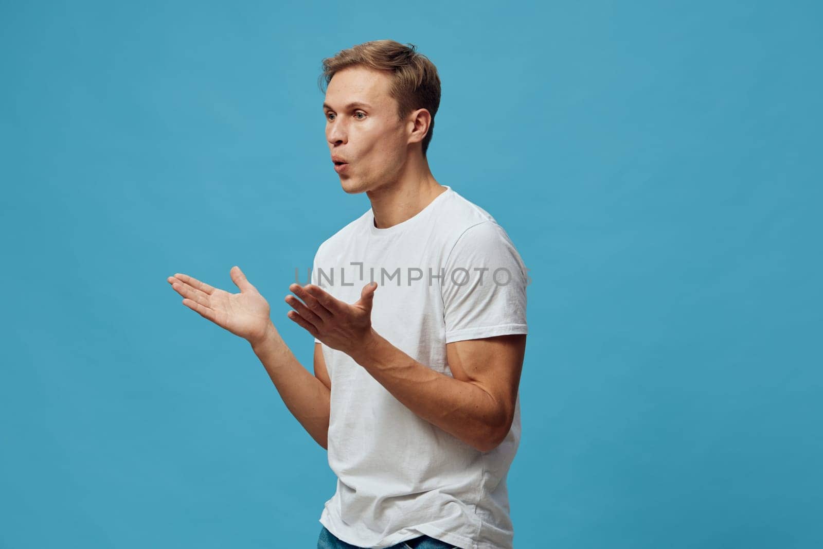 Young man with short hair in casual white t-shirt gesturing with palms facing upward, showing confusion or disbelief against bright blue background, studio shot. People lifestyle concept