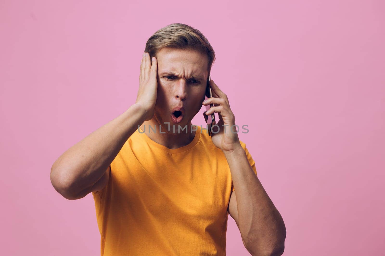 surprised young man in orange t-shirt holding smartphone to ear with shocked facial expression and hand on head against pink background. Concept: digital detox and emotional reaction to smartphone use