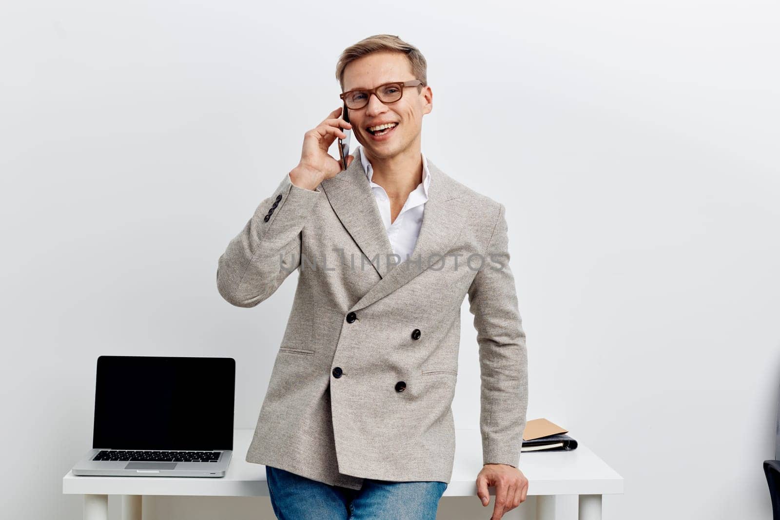 smiling man talking phone wear casual business jacket and glasses standing near laptop on desk in bright room modern workspace remote work digital nomad concept communication connection