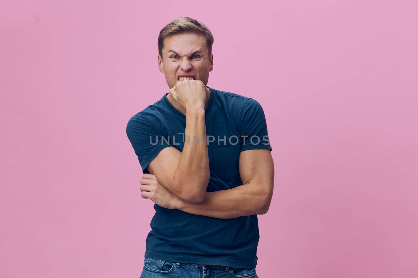Portrait of cheerful man with confident expression, wearing casual blue t-shirt and jeans, standing with arms crossed and hand on chin in front of pink background. Close-up shot capturing joyful, relaxed mood, modern style, lifestyle concept, positive attitude, studio setting.