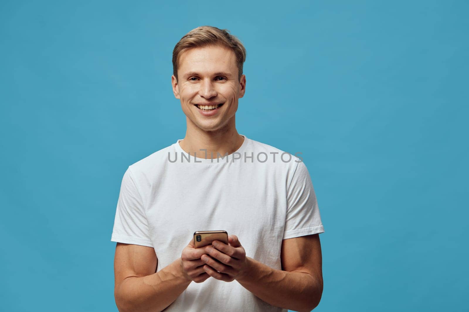 Confident young man with a bright smile holding a mobile phone, standing against plain blue backdrop. Casual outfit and positive vibe highlight modern technology usage, upbeat atmosphere, lifestyle, communication, happiness, connection, studio photography.
