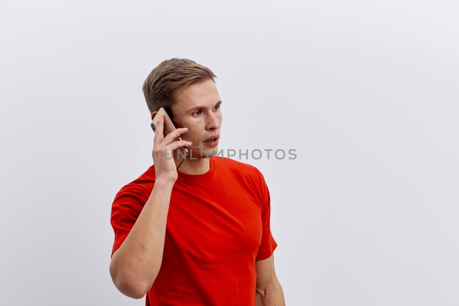 Young caucasian man talking on mobile phone wearing red t-shirt with serious expression isolated on white background. Modern communication and digital nomad lifestyle concept with mobile technology used for global connection.