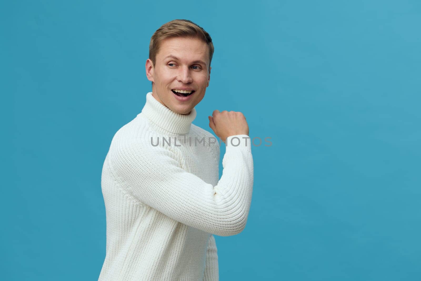 Smiling man in white sweater looking back over shoulder on plain blue background. Young caucasian man expressing joy and confidence in casual wear. Concept: digital nomad lifestyle with remote work and technology.