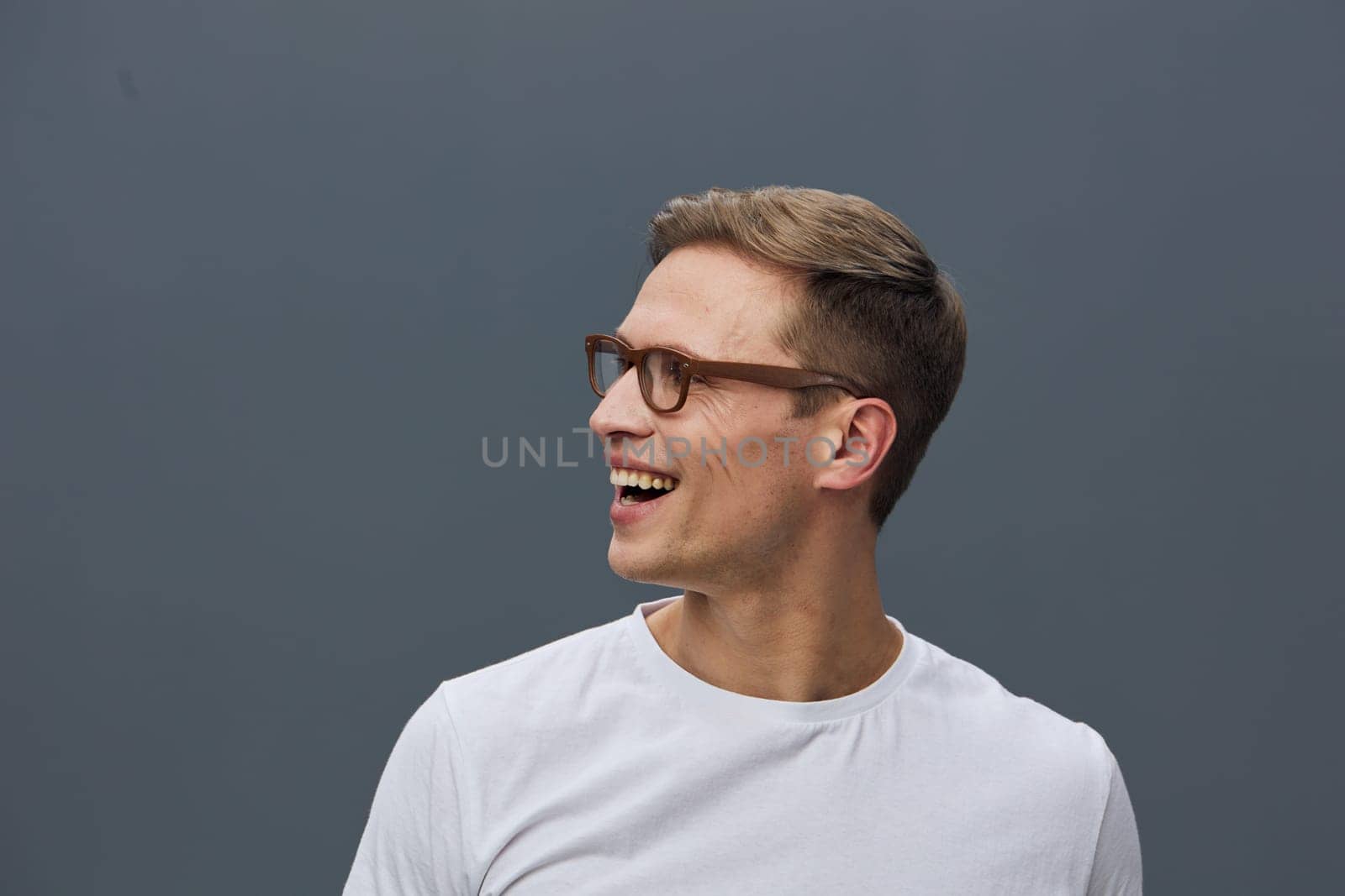 Happy man with short brown hair and glasses wearing a white casual t-shirt, smiling and looking to the side, with a plain gray studio background. Concept: joyful, casual, positive mood.