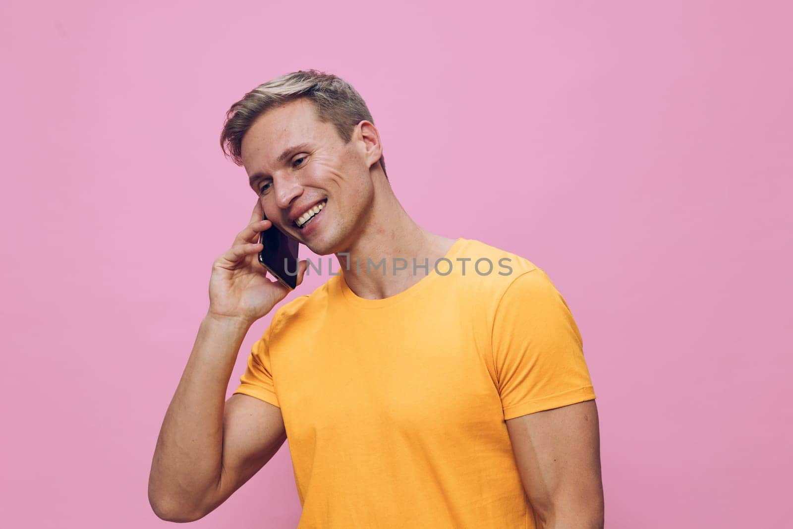 Happy smiling man engaging in a phone conversation on a mobile device, dressed in a vibrant yellow t-shirt, standing in front of a pink backdrop. Modern casual style, youthful energy, positive mood, lifestyle concept, studio setting, colorful scheme, relaxation