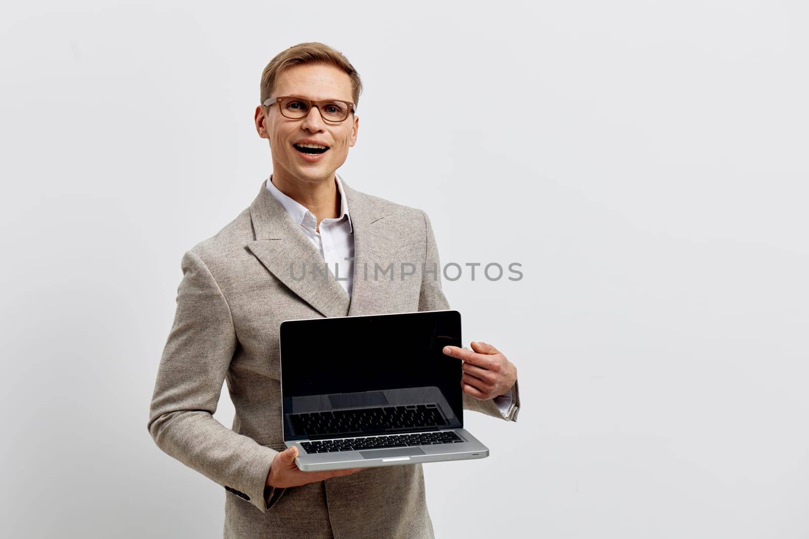 Cheerful man with glasses dressed in a gray blazer holding a laptop computer, standing confidently against a plain white backdrop. Suitable for technology, business, remote work, professional, modern workplace, success, workplace concept